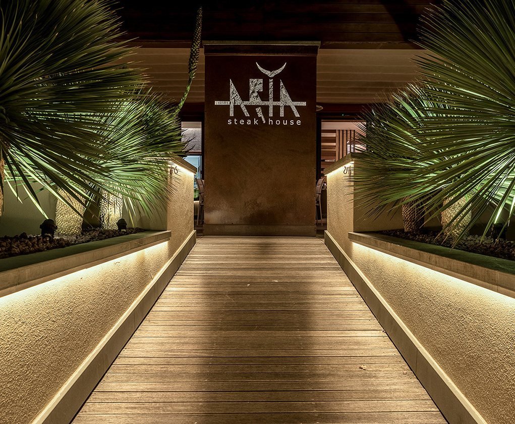 La passerella di legno illuminata tra le palme, verso l’insegna di Aria